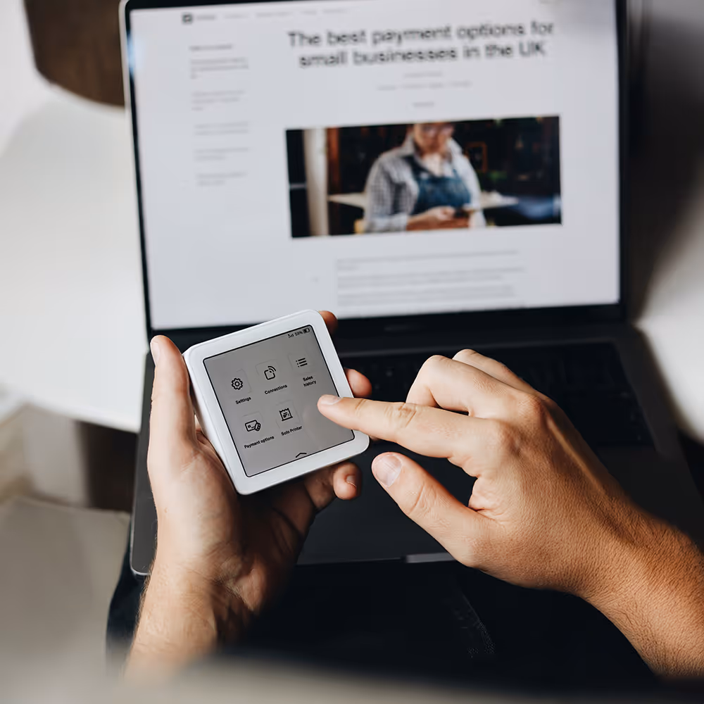 zwei Hände bedienen ein digitales Bezahlsystem, im Bild-Hintergrund ein geöffneter Laptop